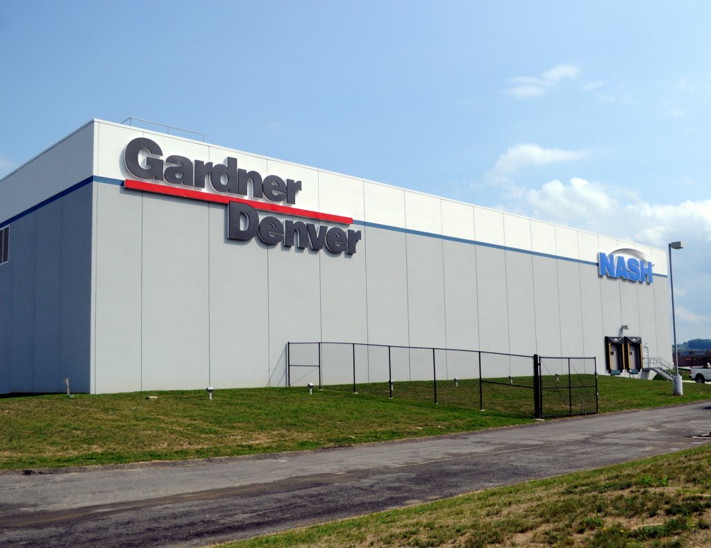 Large industrial building with "Gardner Denver" and "NASH" logos, set against a clear blue sky. Green grass and a metal fence line the path in front.