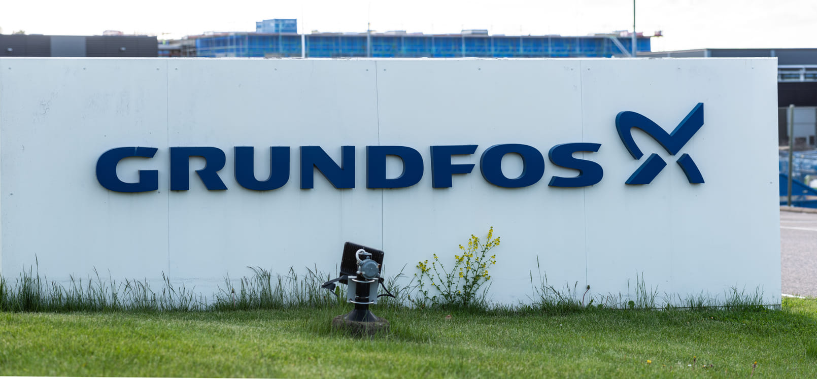 White sign with blue "Grundfos" logo and text in the foreground. Set in a grassy area with industrial buildings and a water pump in the background.