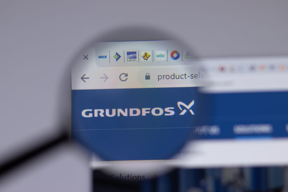 A magnifying glass focuses on a computer screen showing the Grundfos logo. The logo is white on a blue background, with browser tabs visible.