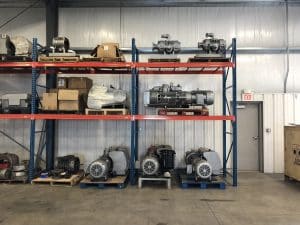 Warehouse interior with metal shelving holding industrial machines and boxes. The setting is organized, with a door and an exit sign visible on the right.