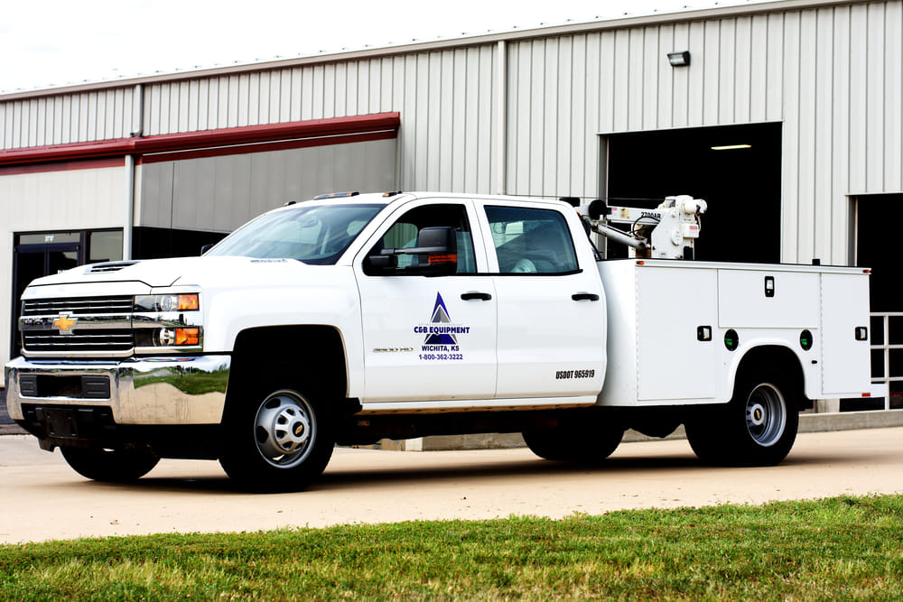 White utility truck with company logo parked in front of a beige warehouse. The truck has a service body and equipment on its bed.