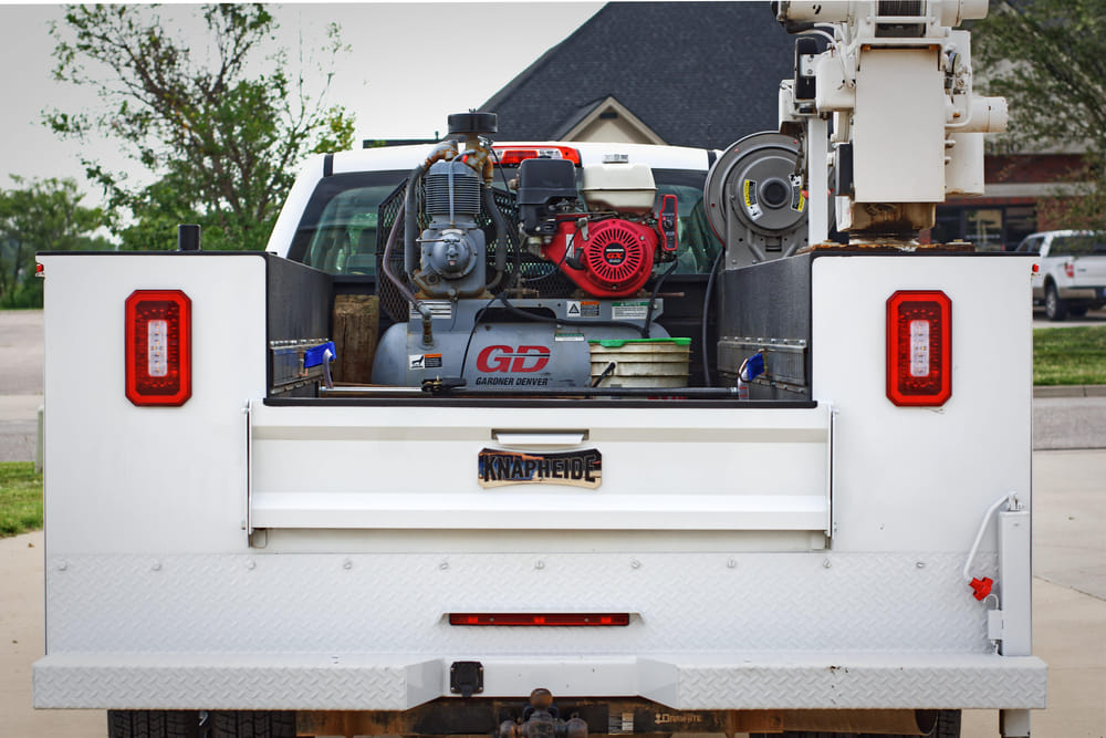 White utility truck with cargo bed open, revealing an air compressor, red engine, and various tools. Overcast sky, suburban background.