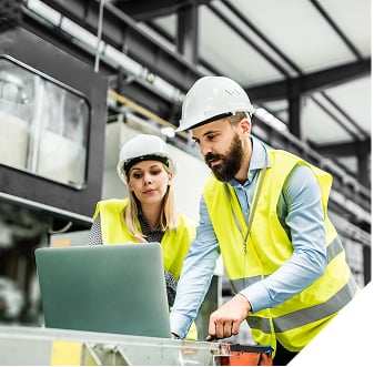 Two workers in safety vests and hard hats review data on a laptop in an industrial setting. They appear focused and engaged in discussion.