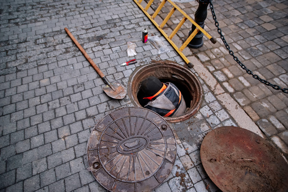 A worker in an orange vest is partially visible inside an open manhole on a cobblestone street, surrounded by tools and a ladder, conveying an active work scene.