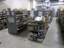 Dimly lit storage room with metal shelves lined with organized stacks of books and boxes. The space feels utilitarian and orderly.
