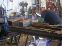 A person in protective gear welds metal pieces in a busy workshop, surrounded by tools and machinery, conveying focus and craftsmanship.